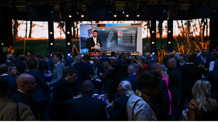 Men in suits, a few women in between, everyone talking, party atmosphere. In the background, a screen on which a moderator presents election results.
