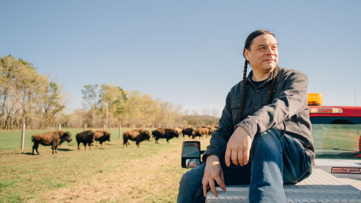 Seah Sherman is sitting on the back of his truck, he has two braided pigtails and is wearing jeans. Behind him, you can see buffalo grazing.