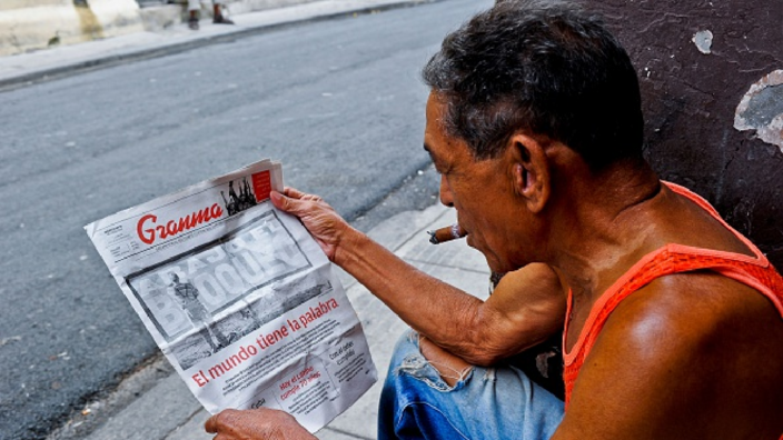 The picture shows the view over the shoulder of a man. He wears a sleeveless shirt and torn jeans and smokes a cigar. He sits in what appears to be a doorway on a street. Over his shoulder we see a newspaper, which he is reading.