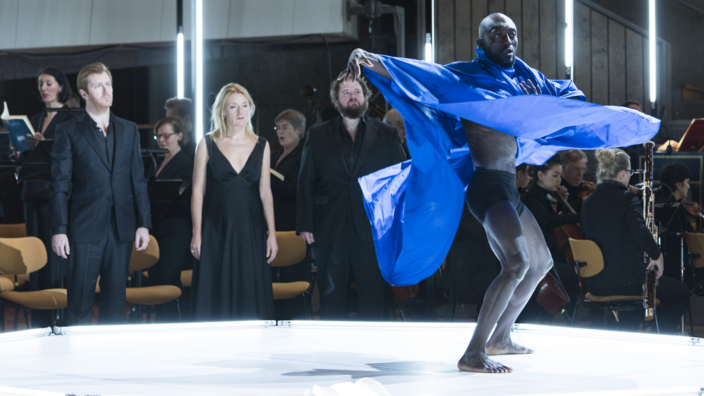 A man with black skin dances on a round white stage. He wears a wide blue cape. At the edge of the stage are two men and a woman in their midst. All of them are dressed in black. In the background is a choir. On the right sits part of the orchestra.
