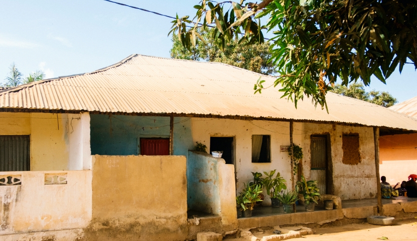 Ein einfaches lehmverputztes Haus mit einem Wellblechdach steht leicht erhöht in Guinea-Bissau in Westafrika. Die Tür ist offen. Vor der Tür stehen Topfpflanzen.