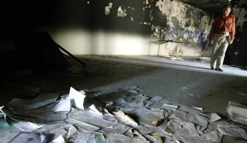 On the floor of a room are scattered many papers and documents. A man with a newspaper under his arm looks at the destruction.