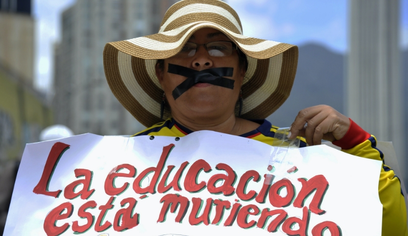 A man with a hat holds up a sign that says "Education is dying" in Spanish. The man has taped his mouth shut with black tape. 