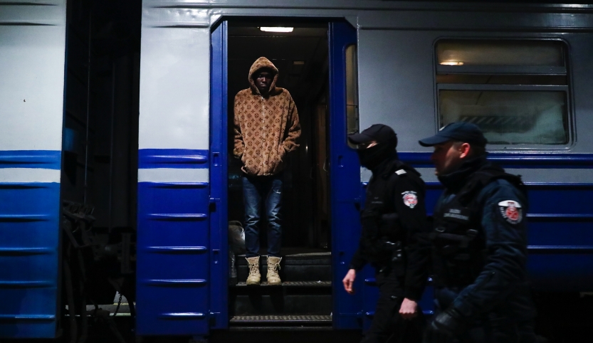 The picture shows a train coach standing in a dimly station. In the open door, a black man is standing and looking onto the platform. Two police men walk by.   