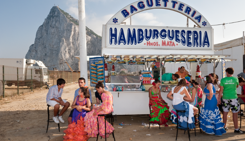 A hamburger stand with a large advertising poster. In front of it, a dozen young people sit on black bar stools. The young women are wearing long colorful voillant dresses, the young men shorts and shirts. Behind a fence is a white truck. In the background is a rocky mountain.