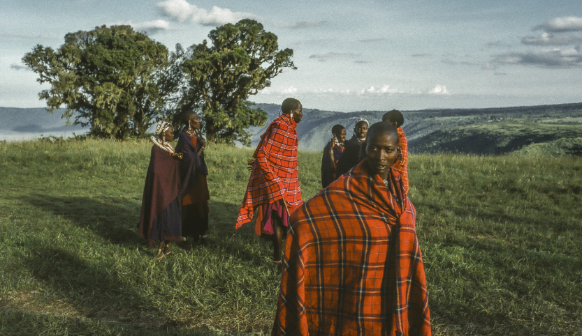 A small group of people with black skin color stands on a green meadow. The part of the group wears red/brown checkered capes, the other part brown capes. One person looks into the camera. In the background is a tree, behind it is a precipice.