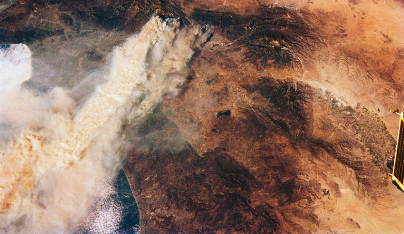 Aerial view of a red-brown landscape, steam rising from a crater.