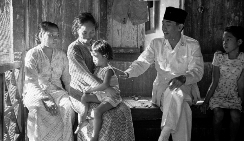 Ein junger indonesischer Mann in einem weißen Anzug in Uniformstil und einer schwarzen Kopfbedeckung sitzt mit einer jungen Frau und zwei Mädchen auf einer Bank in einem Haus. Der Mann legt seine rechte Hand auf den Rücken eines ca. drei oder vierjährigen Mädchens, das rechts von ihm auf dem Schoss der Frau sitzt. Der Mann lächelt in die Richtung des kleineren Mädchens. Die Frau lächelt zum Mädchen auf ihrem Schoss. Das Mädchen links von dem Mann ist ca. 10 Jahre alt. Sie sieht in die Richtung der Frau mit dem Mädchen auf ihrem Schoß.  Die Frau und die Mädchen tragen leichte Kleidung. Das Holzfenster ist geöffnet. 
