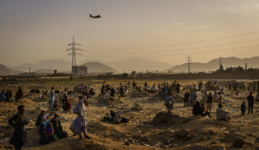 Many Afghan people are standing on a strip of land. In the background are mountains, a few houses and electricity pylons. An airplane flies over the people. 