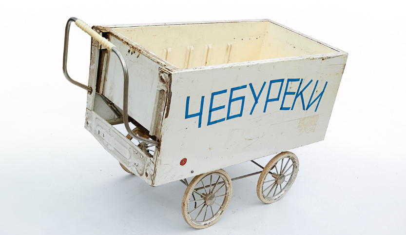 Auf dem Gestell eines alten Kinderwagens liegt ein weißer Kühlschrank ohne Tür mit der Öffnung nach oben. Auf der Seite stehen folgende Ziffern 4E6YPEKU.
