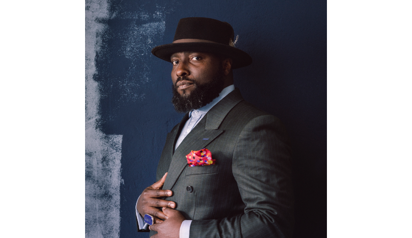 A side portrait of a young black man with a full beard. He wears a gray suit, white shirt and a black hat with brown trim. His hands are resting on his stomach and lapels. On his small right finger is a large blue ring.