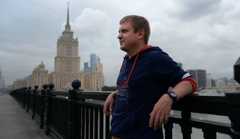 The author Sergei Lebedev is standing on a bridge. He is leaning with his back to the bridge railing. In the background is a high-rise building with several towers in the socialist classicist style.