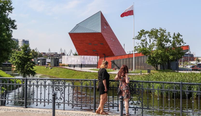 Zwei junge Frauen stehen am Geländer einer Brücke. Sie schauen ins Wasser. Im Hintergrund sind drei moderne Gebäude in rot und orange, davor die polnische Flagge.