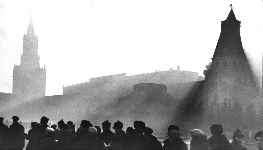 Ein historisches schwarz/weiß Bild. Einige Menschen stehen in einer Stadt vor einem großen Gebäude und zwei Türmen.