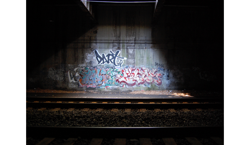 Ein dunkler Raum mit einer Graffiti Mauer, die von einem Licht beschienen wird. Davor verläuft ein Gleis.